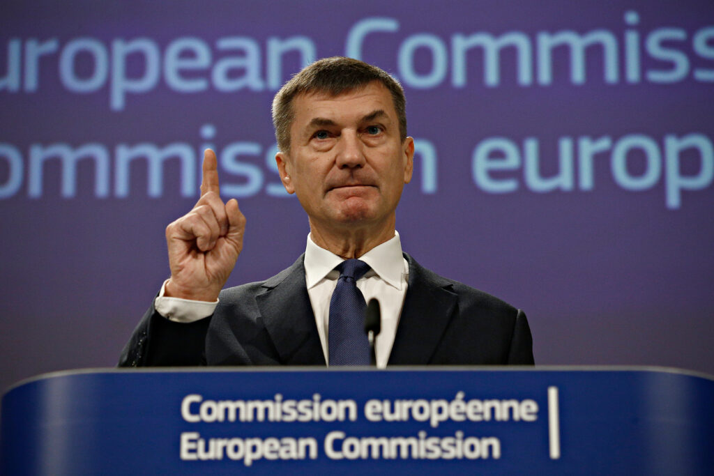Foto Euroopa Komisjoni endisest asepresidendist Andrus Ansipist, kes viibutab oma nipetissõrme. Foto on tehtud Euroopa Komisjonis, Ansip seisab poodiumi ees, seljas on musta värvi pintsak, valge triiksärk ja tumesinine lips.
