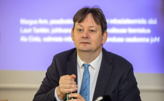 Fotol on riigi valimisteenistuse juht Arne Koitmäe, kes kannab musta ülikonda, valget triiksärki ja sinist lipsu. Koitmäe käes on veepudel. Isik istub laua taga ja tema ees on näha Kanal2 logoga mikrofon.